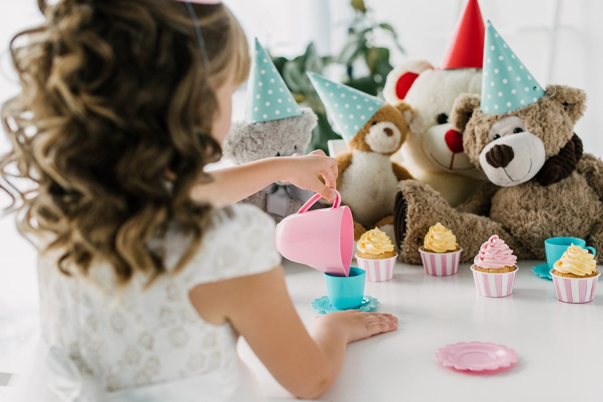 rear view of birthday kid having tea party with teddy bears in party hats at table with cupcakes