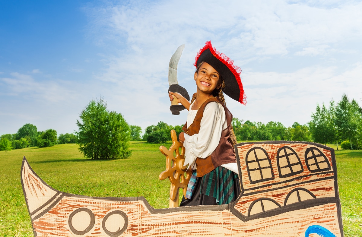 Happy Black girl in pirate costume with hat and sword holds helm on cardboard box ship