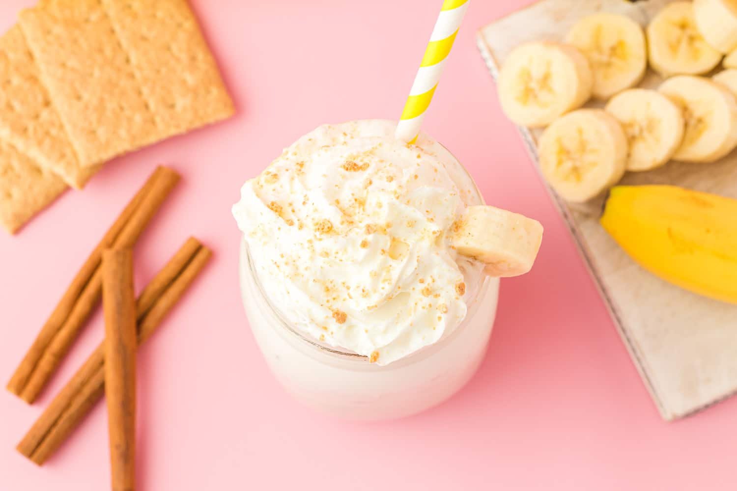 Overhead view of banana cream pie smoothie on pink background with sliced bananas, graham crackers, and cinnamon sticks