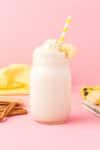 Banana cream pie smoothie (protein shake) in a mason jar on pink background with bananas, graham crackers, and cinnamon sticks