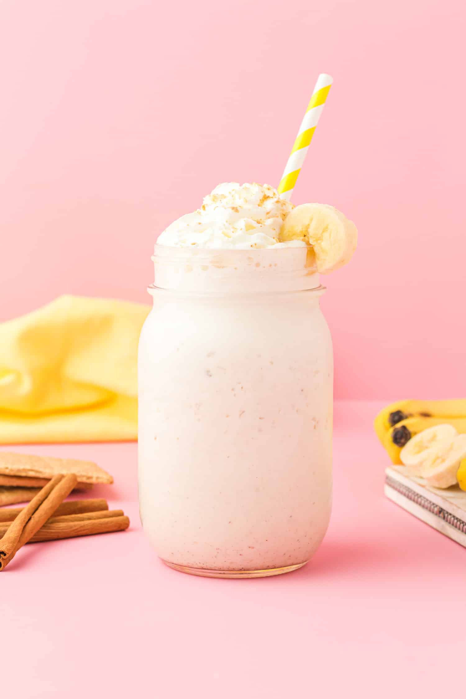 Banana cream pie smoothie (protein shake) in a mason jar on pink background with bananas, graham crackers, and cinnamon sticks