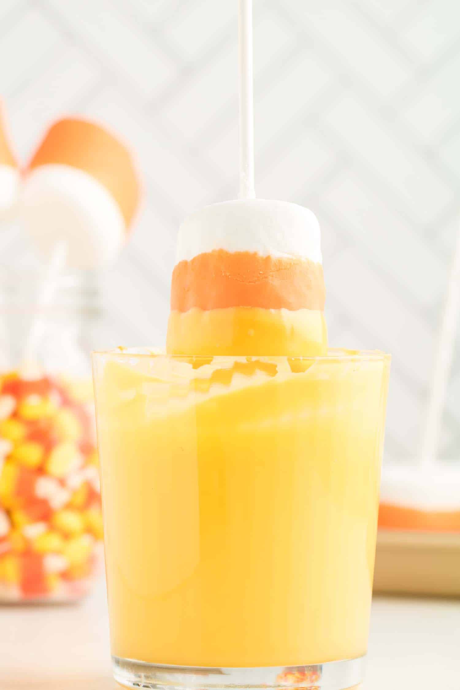 Candy corn marshmallow pops being dunked into yellow candy melts in a glass.
