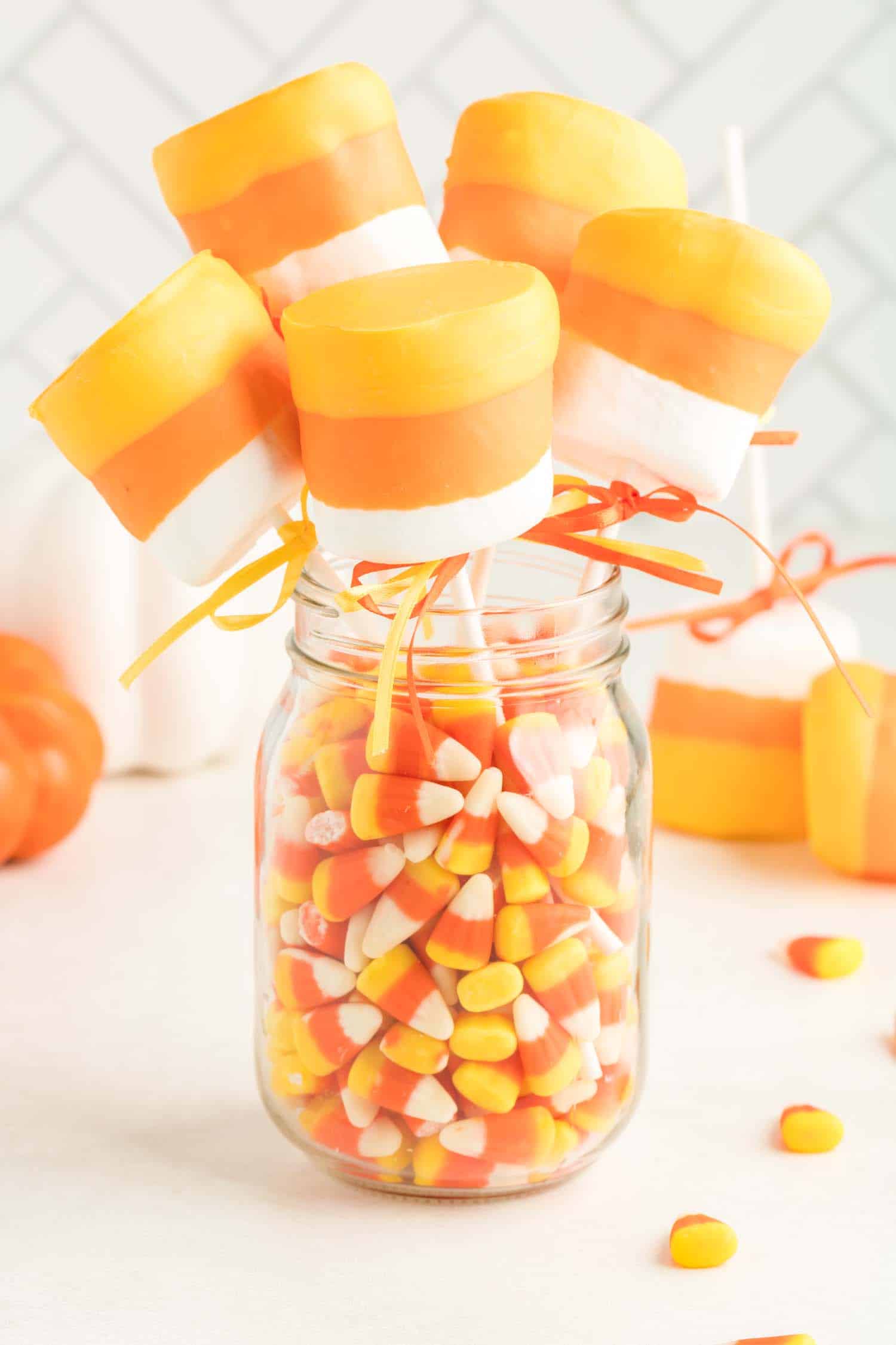 Five candy corn marshmallow pops in a glass jar filled with candycorn.