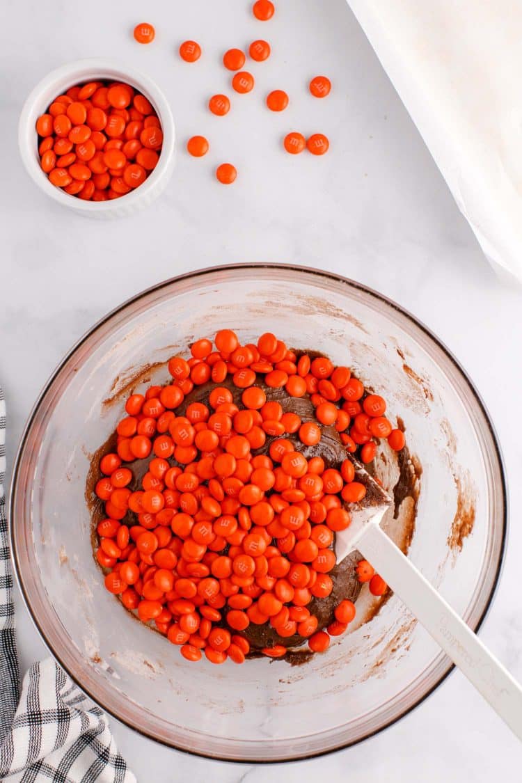 Adding in orange M&Ms to the chocolate cake mix in a glass bowl.