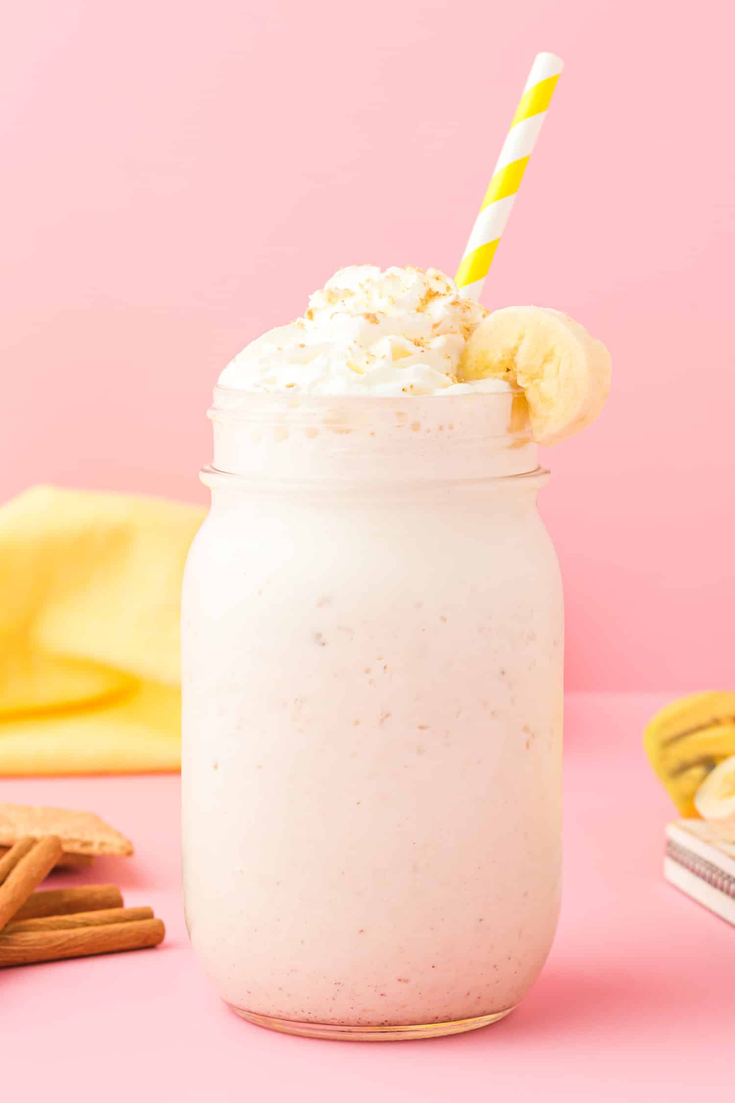 Banana cream pie smoothie in a mason jar glass with yellow and white straw on pink background