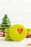 Grinch Stained Glass Cookie propped against stack of green cookies with Christmas tree background