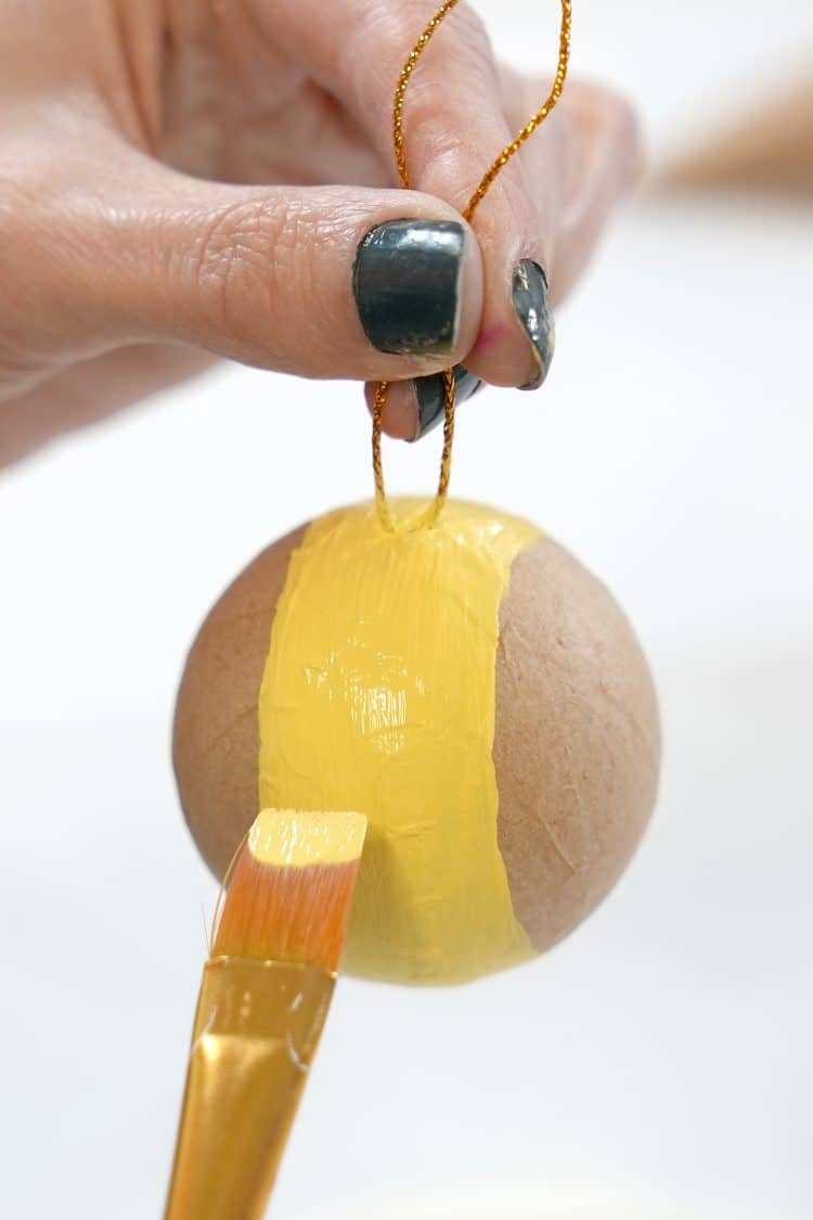 Close-up of hands painting a round kraft paper ornament with a stripe of yellow paint