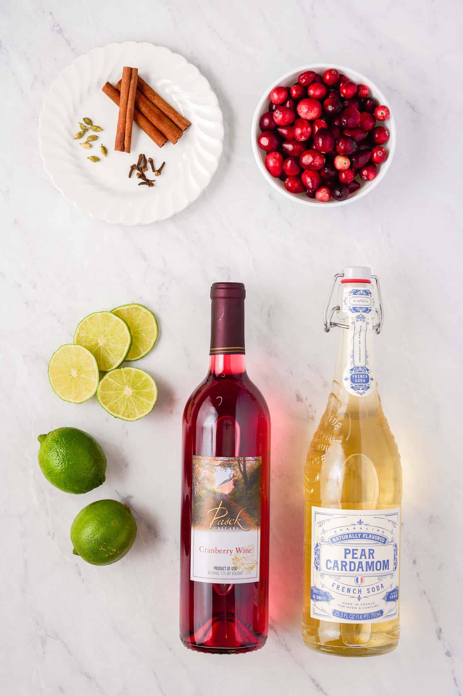 Two wine bottles, limes, cinnamon, and a bowl of cranberries on a white table