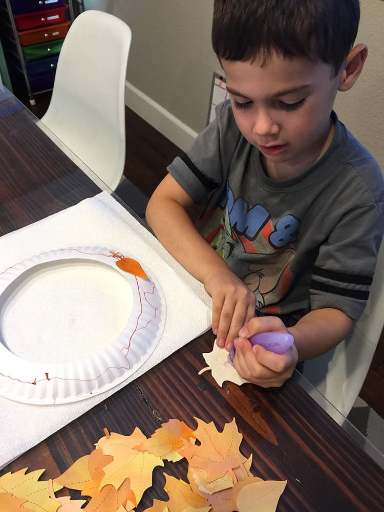 child squeezing glue on the back of leaves with paper plate wreath backing