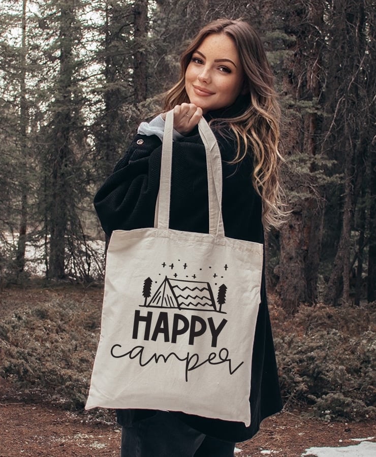 Pretty woman holding a canvas tote bag with "Happy Camper" design in the woods