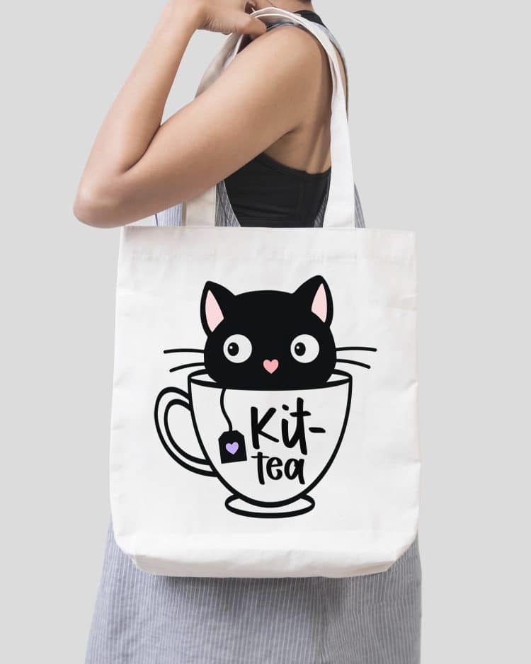 Woman holding a tote bag with a "Kit-Tea" design featuring a black kitten in a teacup
