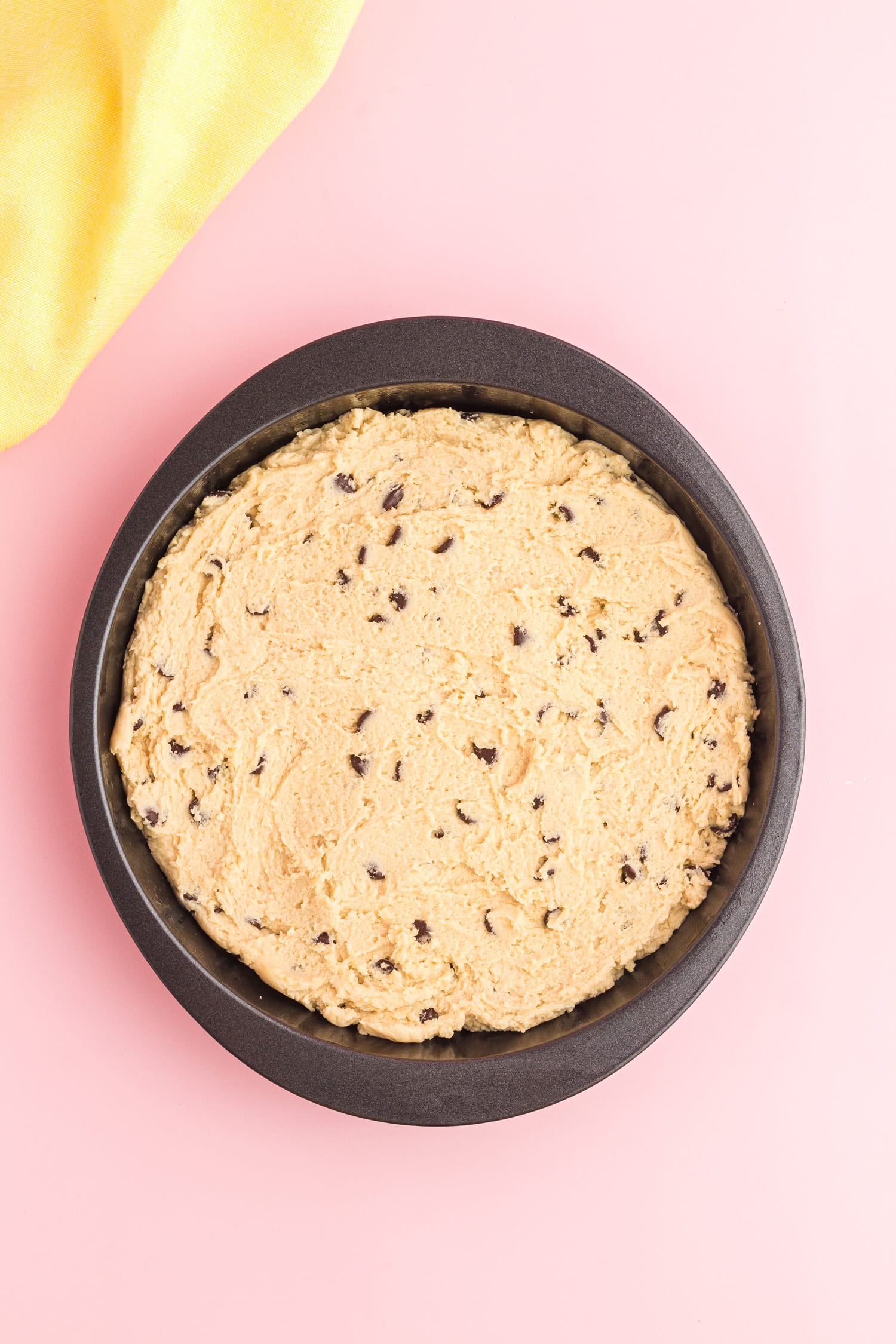 Chocolate chips being added into chocolate chip cookie cake recipe, in a baking dish on pink plain background