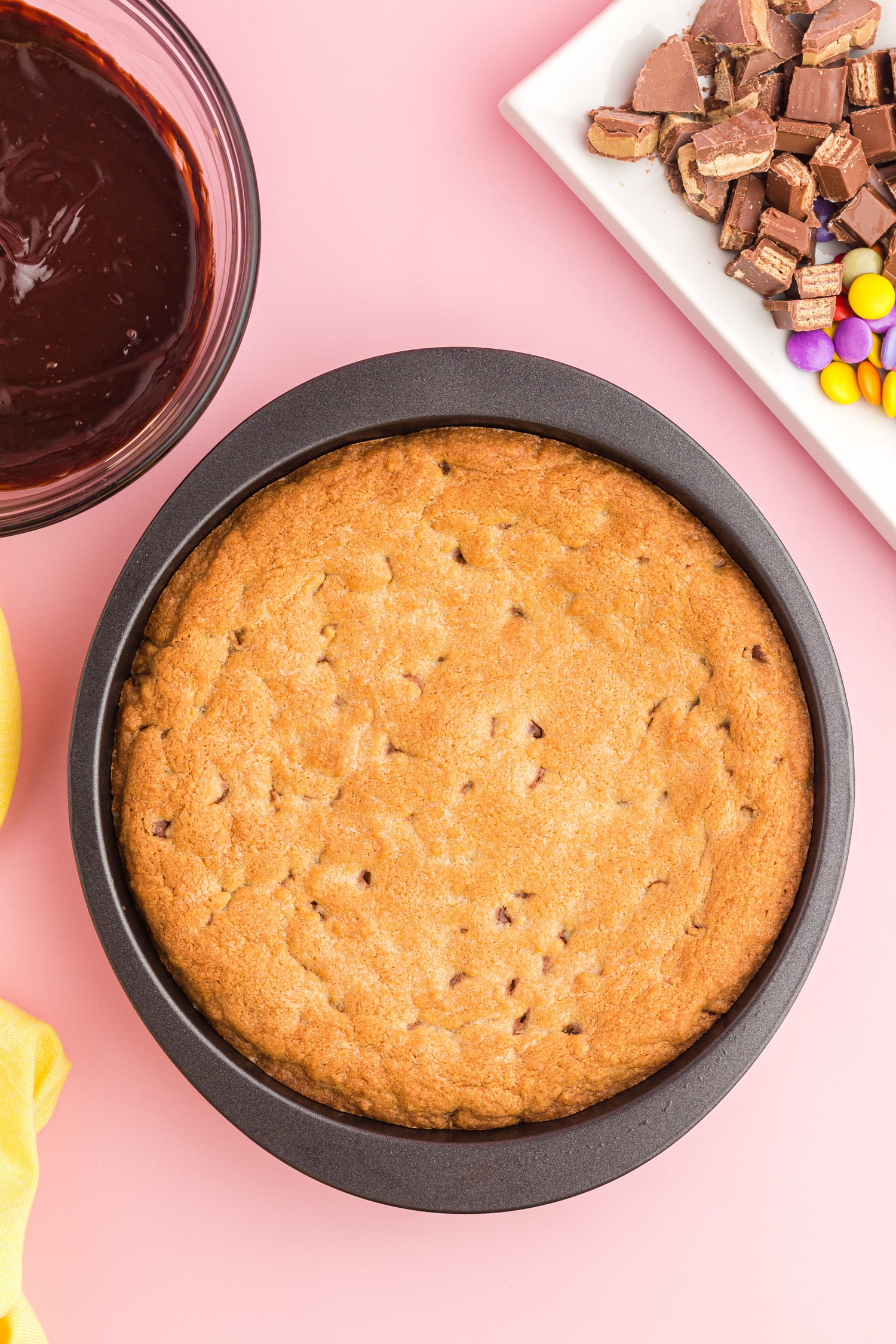 Baked cookie cake cooling on a pink table with candy pictured off in the corner and chocolate ganache topping