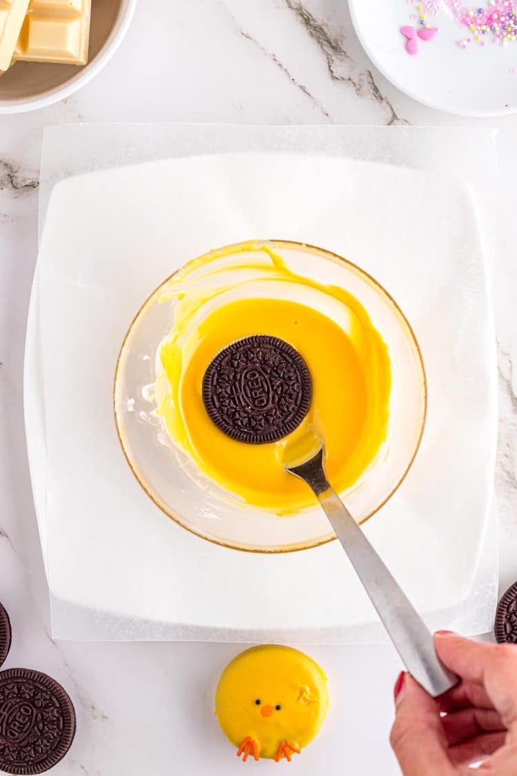 Plain Oreo cookie being lowered on fork into a mixing bowl filled with yellow colored melted chocolate