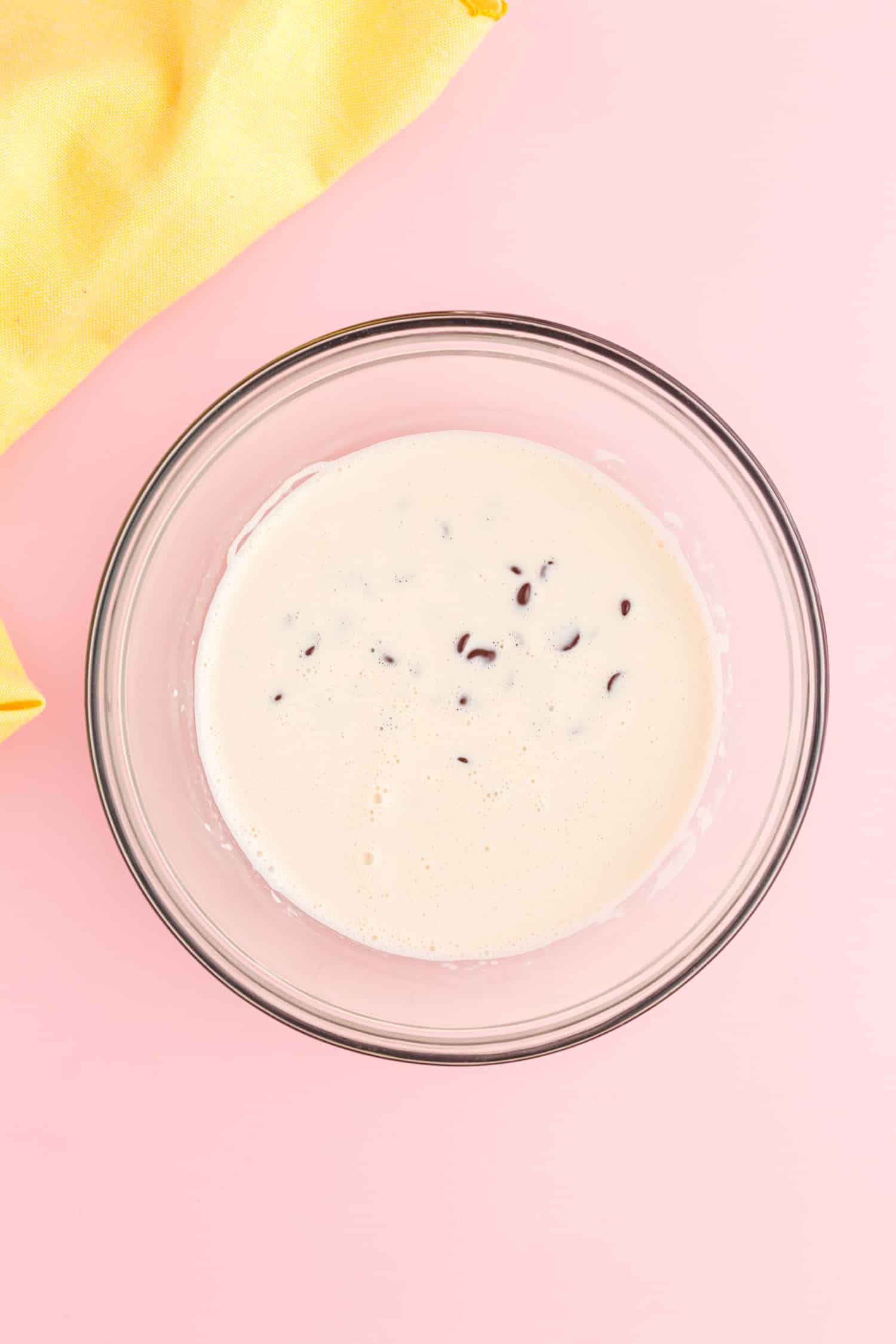 chocolate chips and whipping cream being mixed in clear mixing bowl on pink background