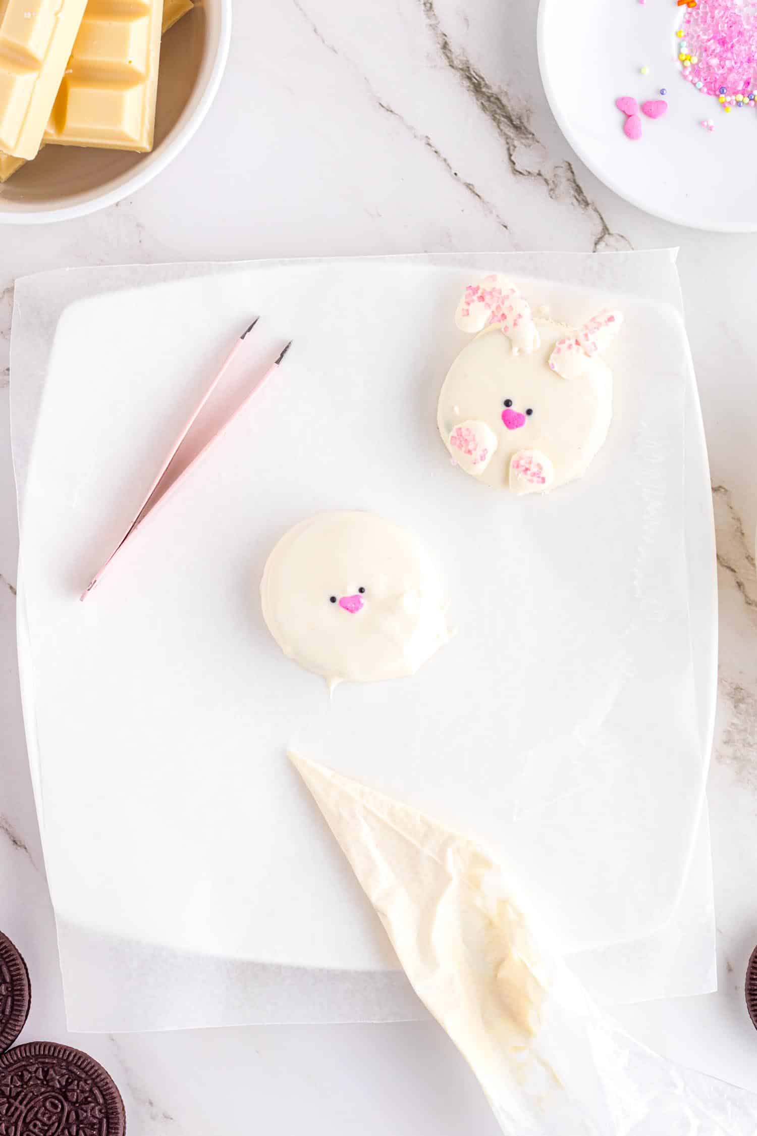 White chocolate covered Oreo cookies being decorated to look like Bunnies using piping bag, tweezers, and sprinkles.