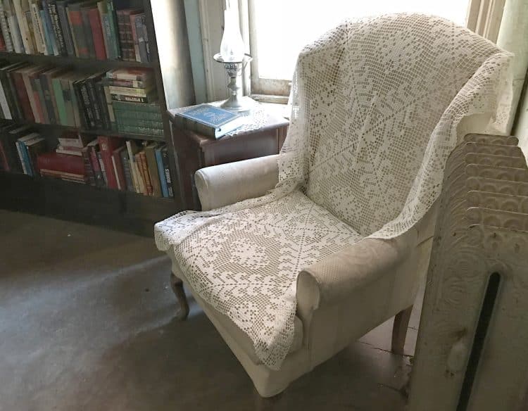 wingback chair covered in large doily next to an overflowing bookshelf