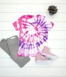 classic spiral tie-dye shirt in shades of pink and purple with shoes and jeans on white wood background