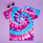 Pink, purple and blue tie dye spiral shirt on a purple background with sunglasses