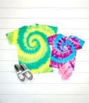 Two spiral tie-dye shirts in shades of yellow, green, pink, and purple, on a white wood background with shoes