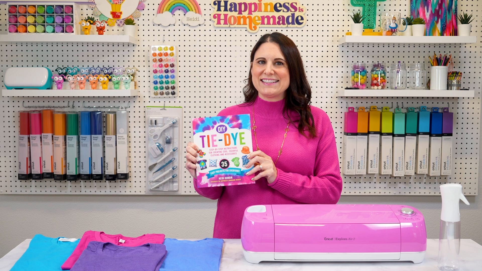 Dark haired white woman holding up "DIY Tie-Dye" book in front of a colorful pegboard wall of craft supplies
