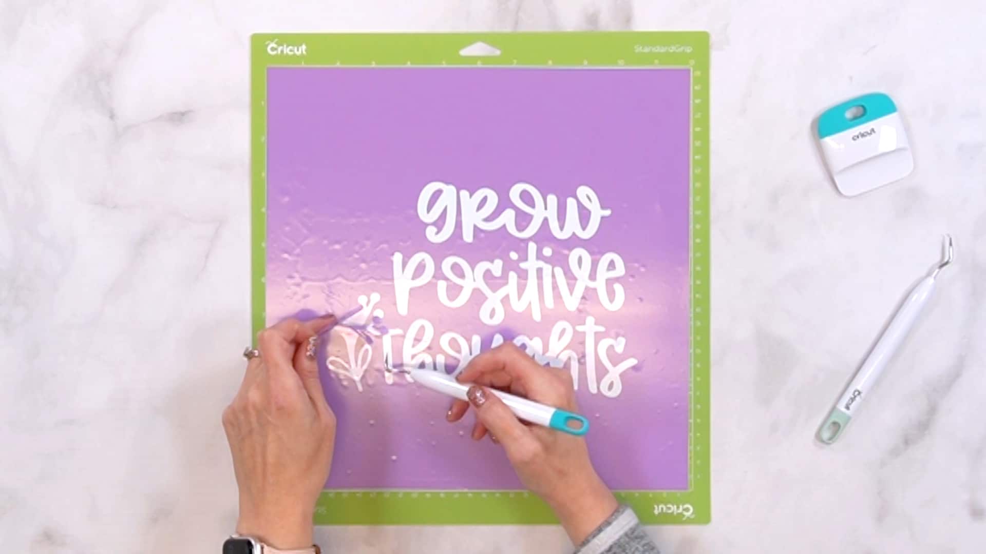 Hands using weeding tools to weed purple vinyl on a green Cricut mat