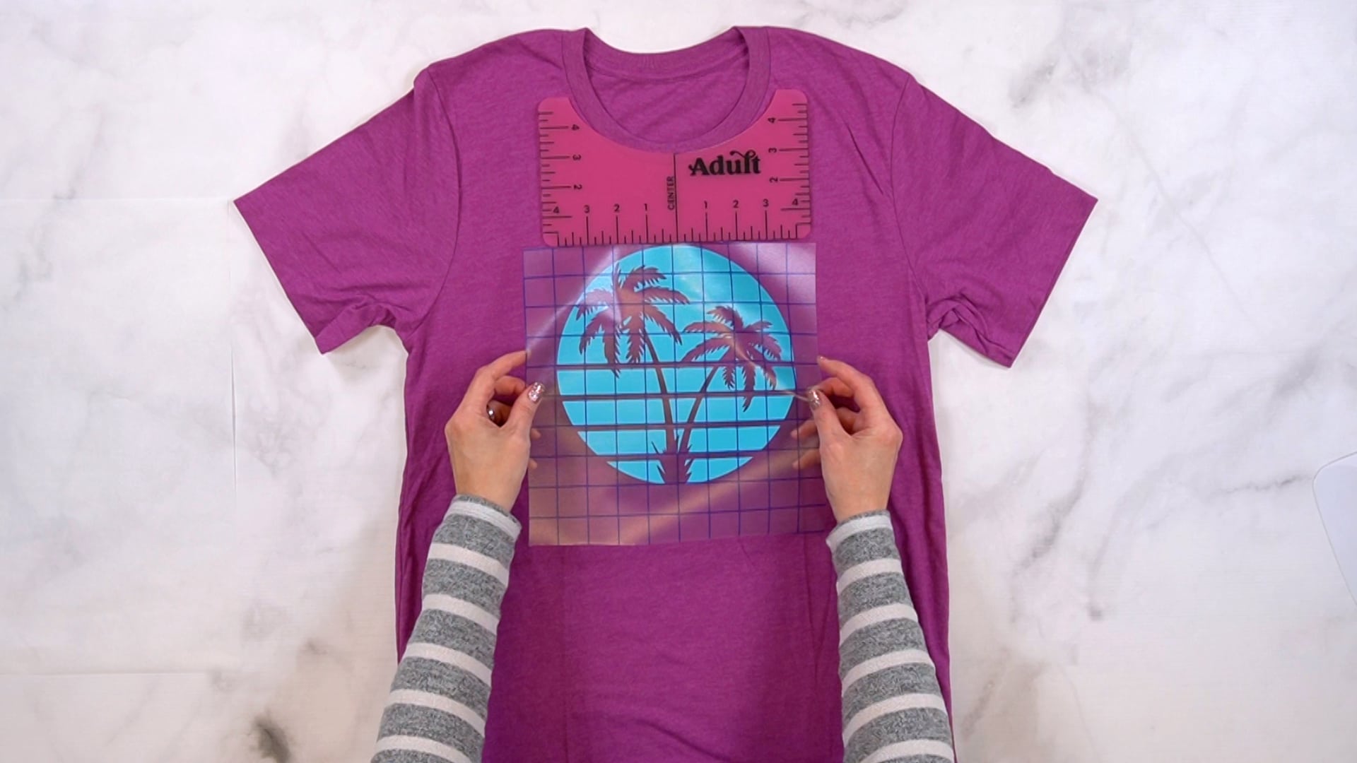 hands positioning blue vinyl sunset and palm tree design on magenta shirt