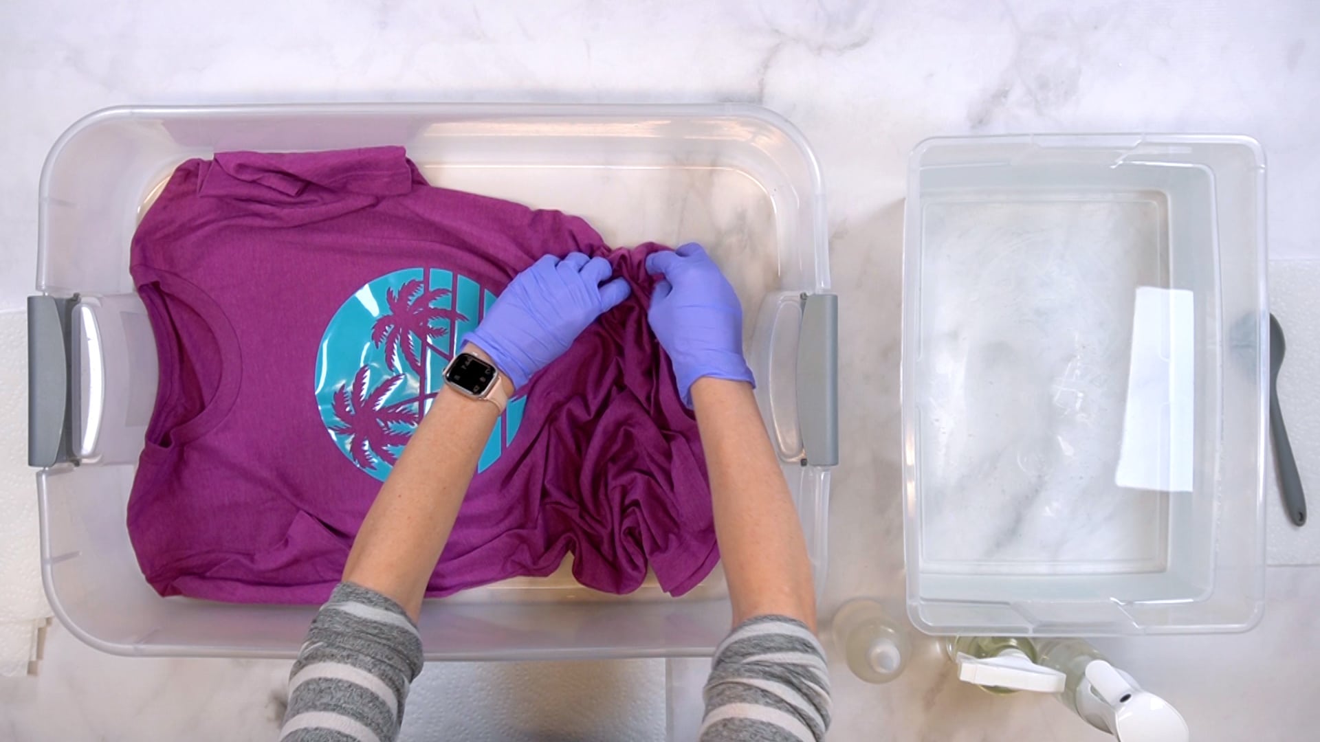 Hands wearing purple gloves scrunching up a purple shirt in a plastic tub