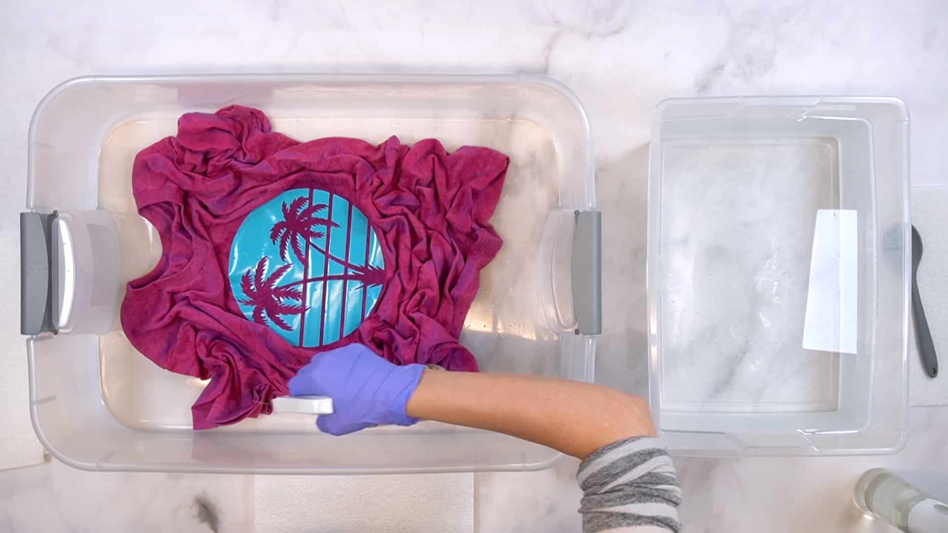 Hands wearing purple gloves spraying bleach solution on a partially bleached magenta shirt in a plastic tub