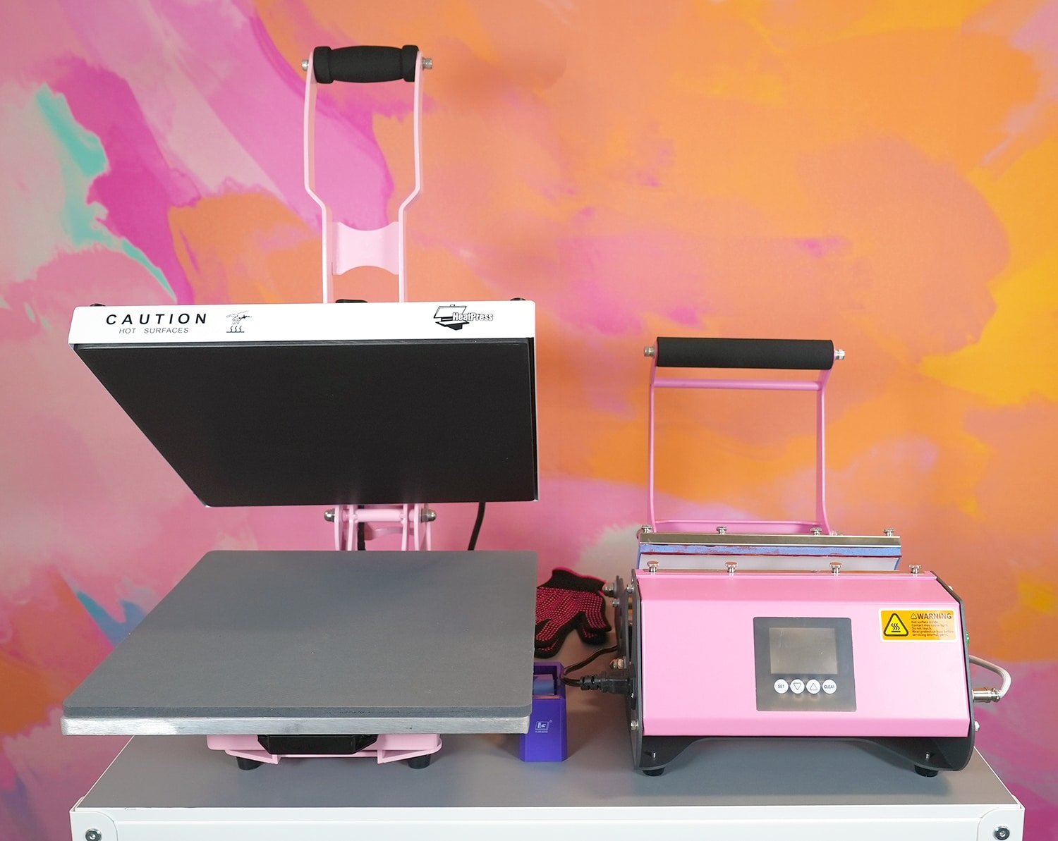 Pink heat press and pink tumbler heat press sitting on a white cart in front of a colorful wall
