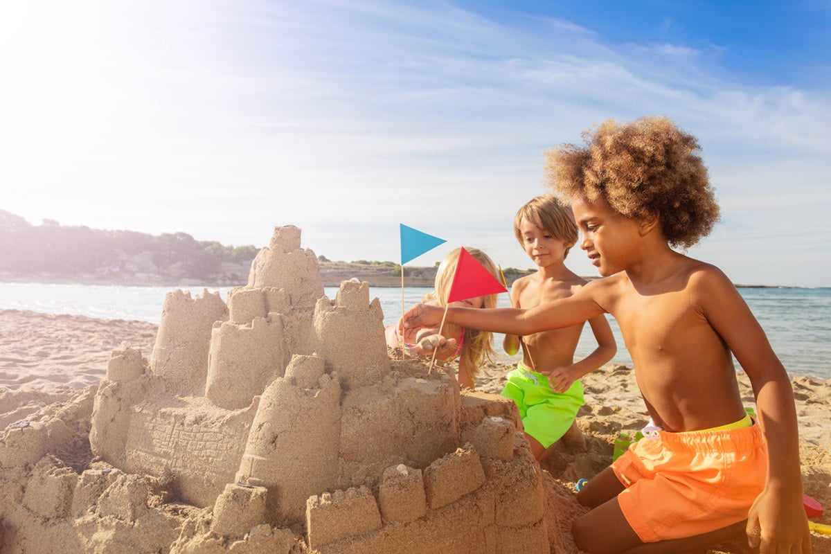 Happy friends, multiethnic boys and girls, playing at the seaside, decorating sandcastle towers with flags