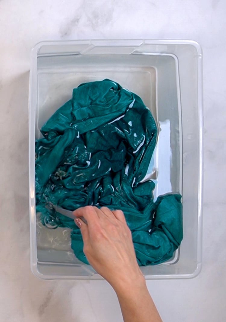 Hand rinsing a teal colored shirt in a tub of bleach neutralizing solution