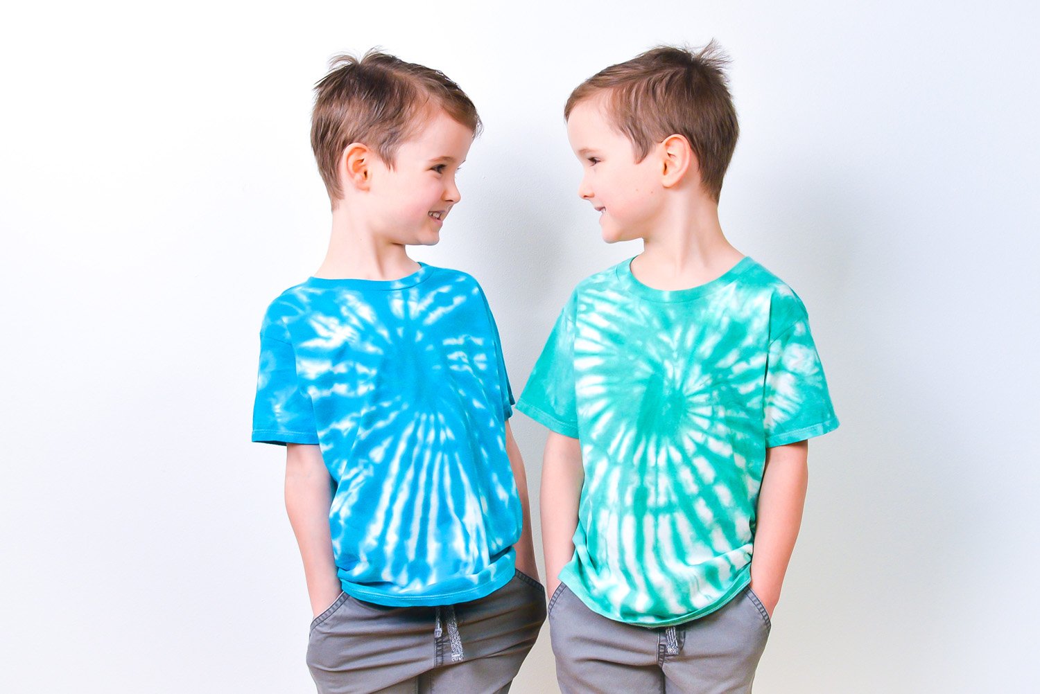 Two 5-year-old twin boys in green and blue tie-dye shirts looking at one another like mirror images