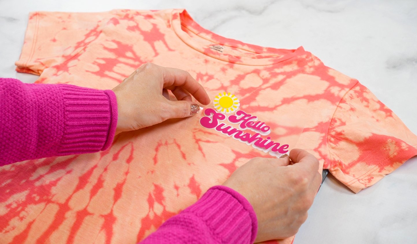 Close up of hands adding a "Hello Sunshine" HTV graphic to a peach and coral tie-dyed shirt