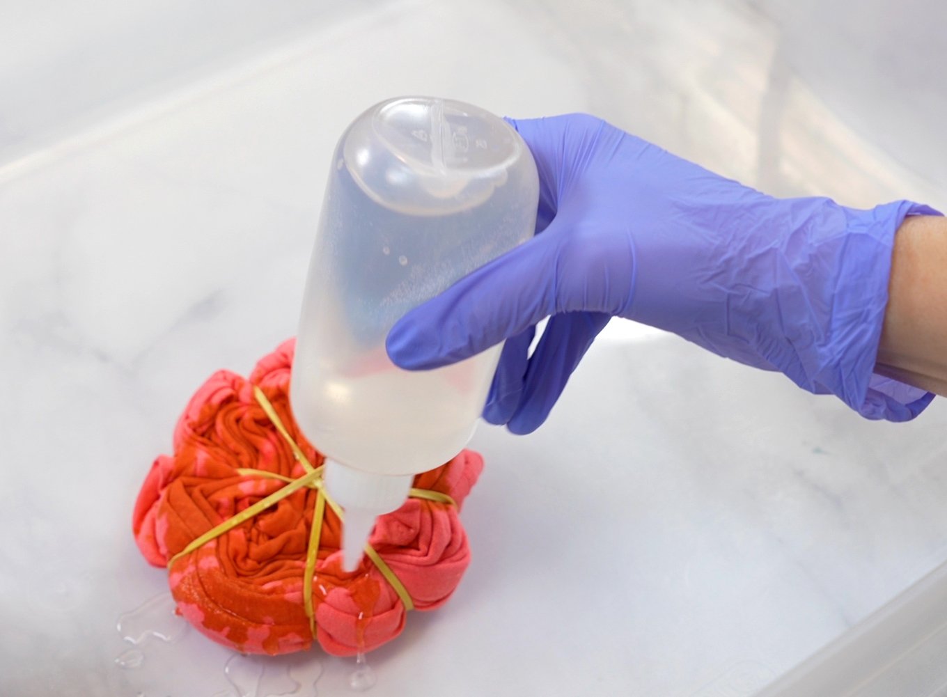 Close up of purple gloved hand holding a squirt bottle of bleach solution that is being squeezed on to an orange shirt