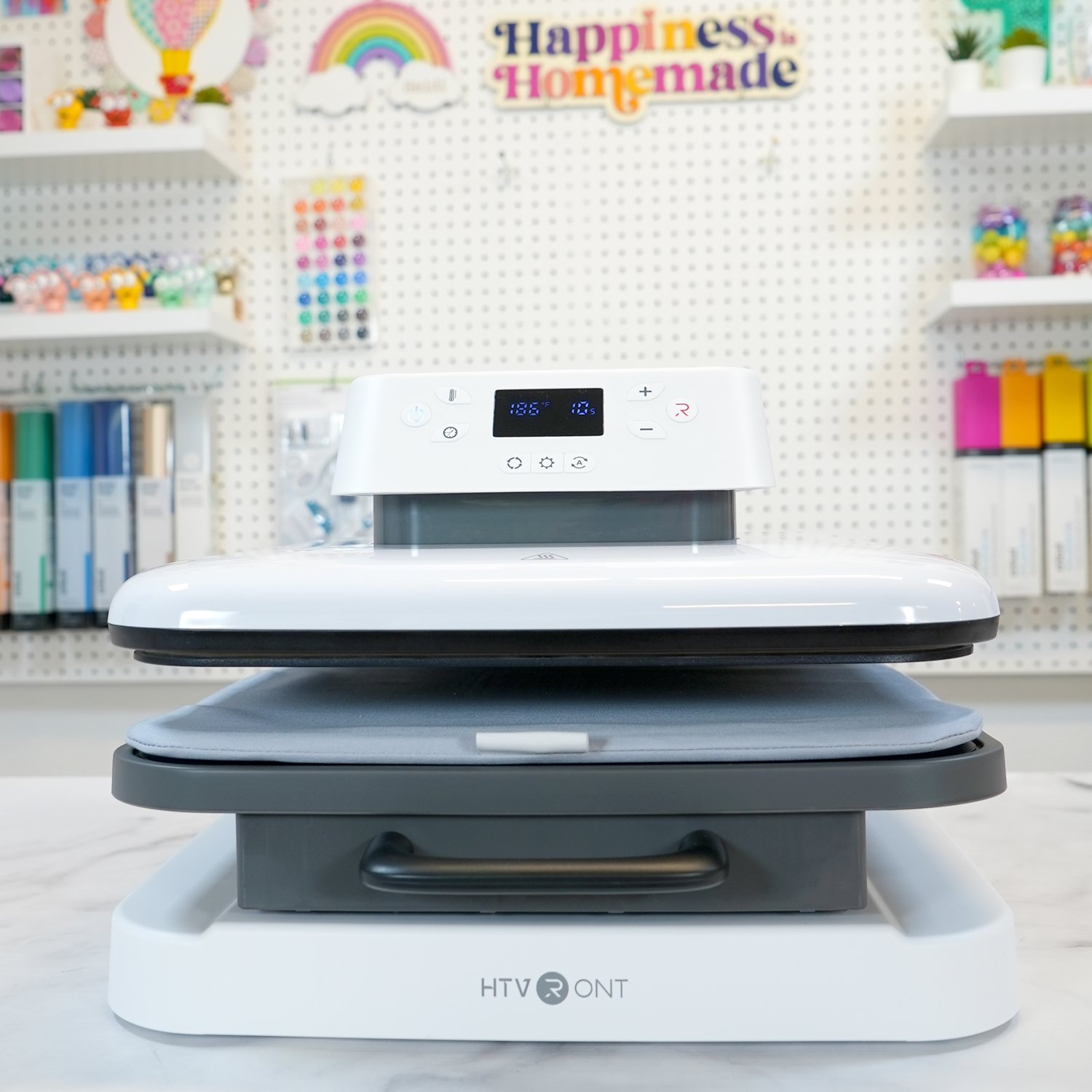 HTV Auto Heat Press on counter in front of colorful pegboard of craft supplies