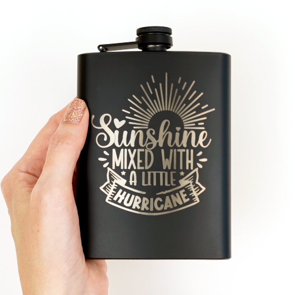 Hand holding an engraved black flask with "sunshine mixed with a little hurricane" design