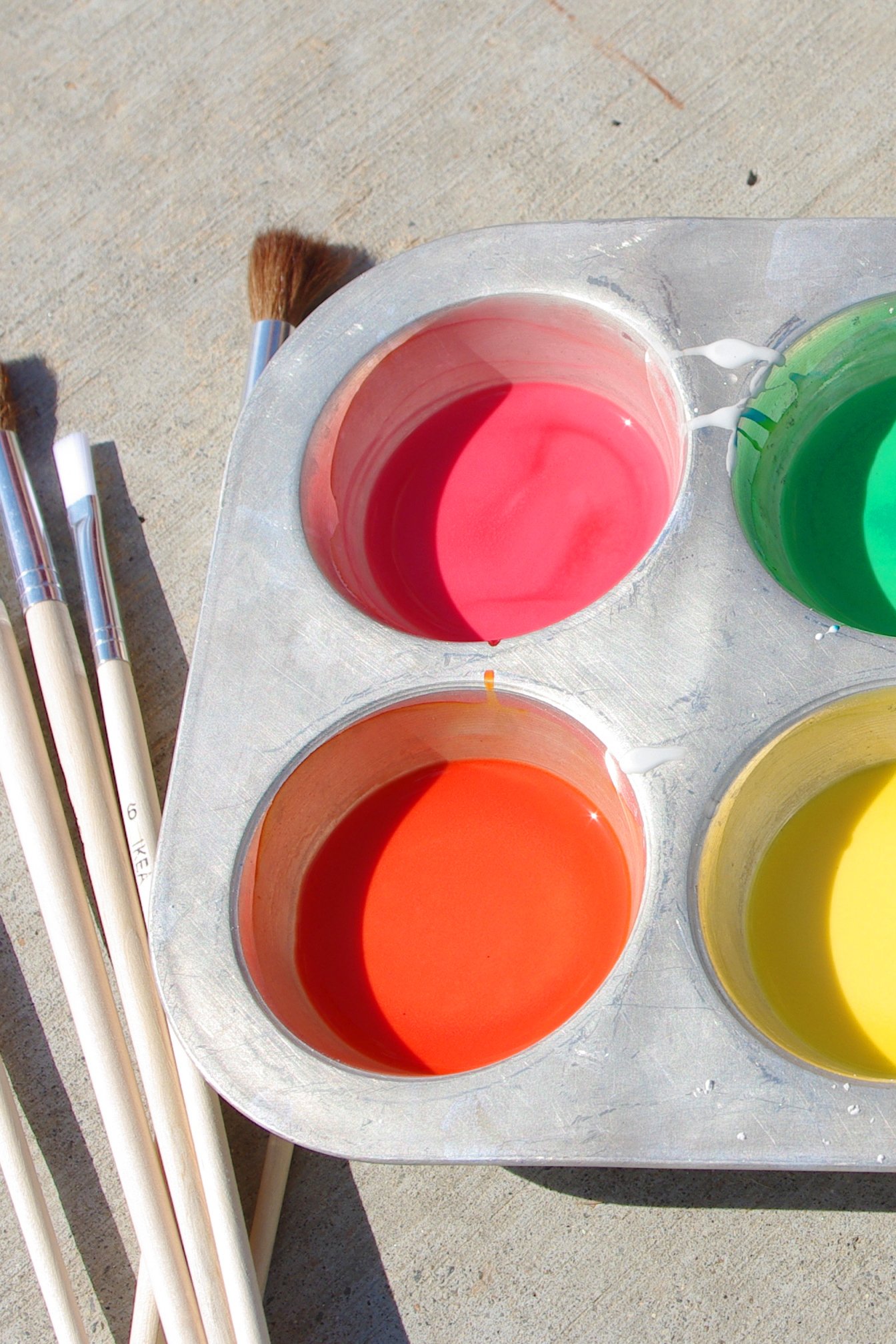 Muffin tin filled with colorful sidewalk chalk paint on sidewalk next to paintbrushes