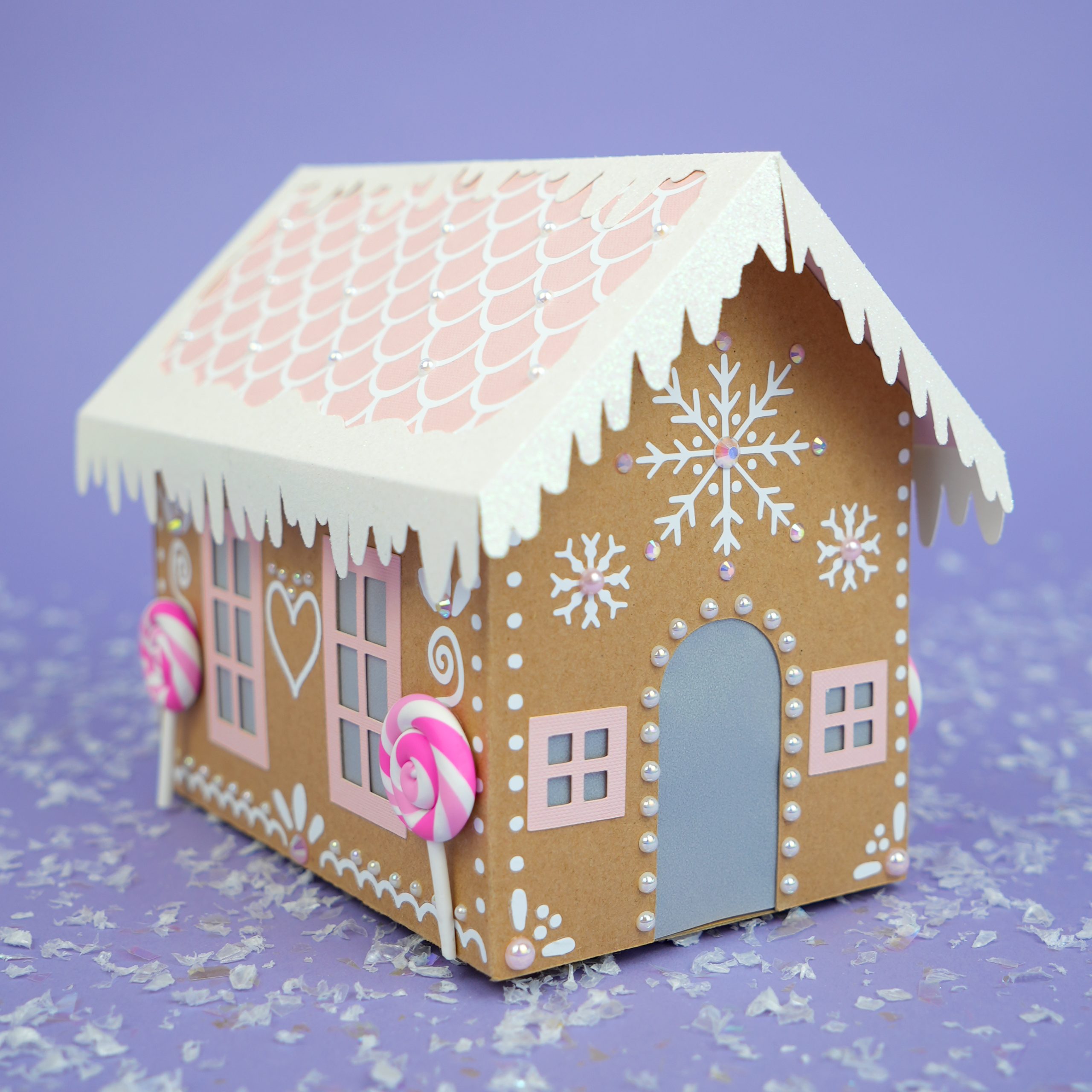 Pastel pink and white decorated paper gingerbread house with pearls and rhinestones on lavender background with faux snow