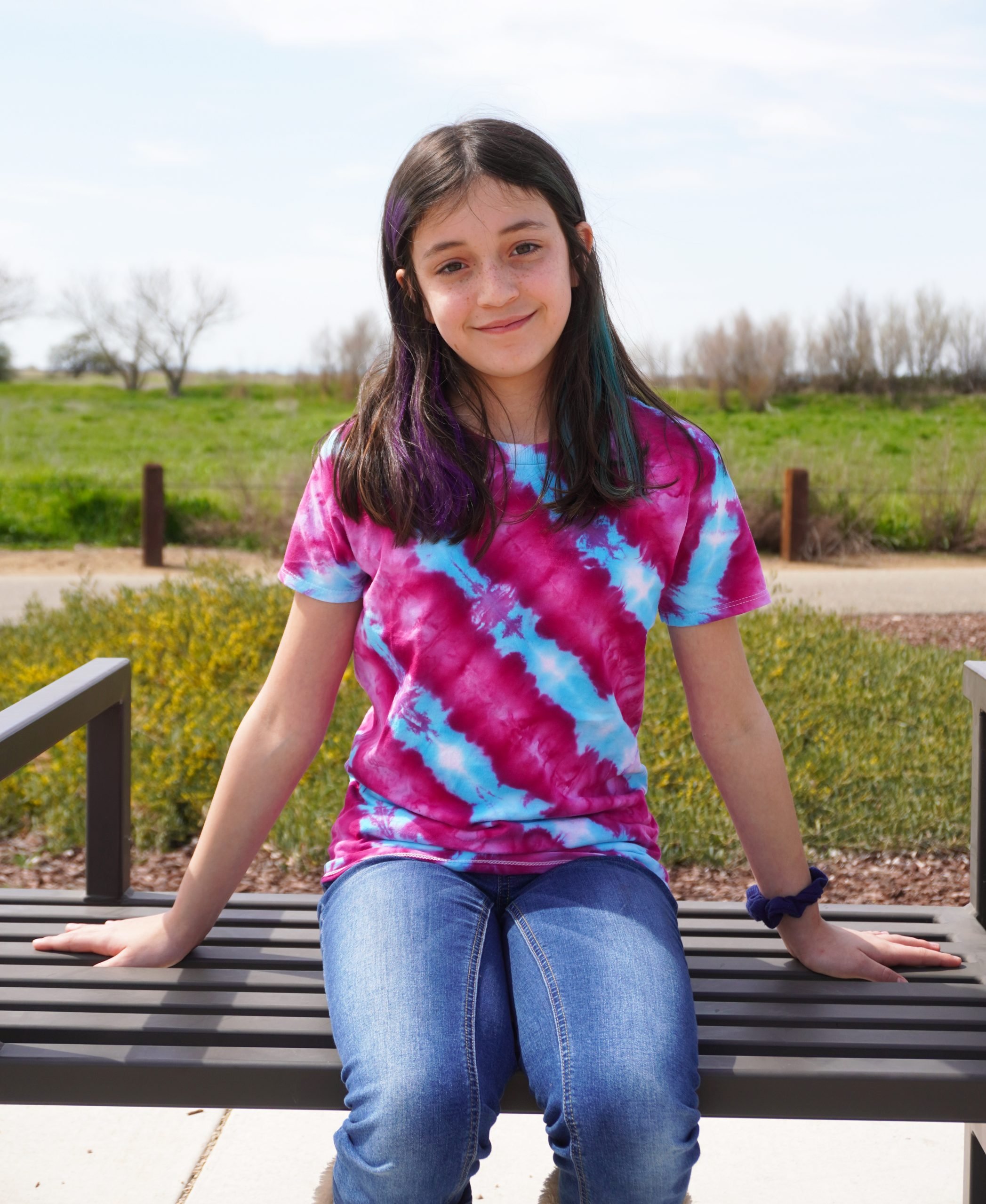 Dark haired 12 year old girl wearing a pink and blue tie dyed t-shirt on an outdoor bench