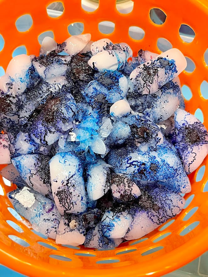 Pile of ice with blue dye powder on top of an ice dye t-shirt inside a plastic basket with holes