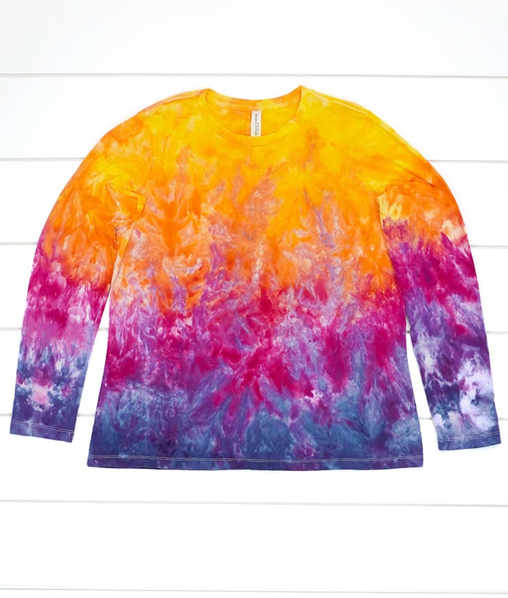 Ice dyed long sleeve t-shirt in sunset shades of yellow, orange, pink, purple, and navy blue on white wood background