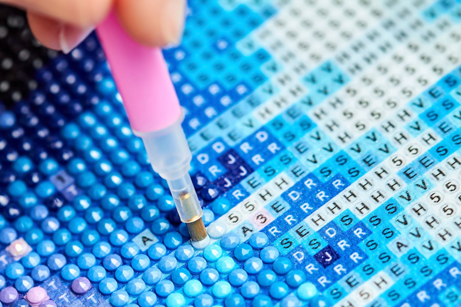 Close up of pink diamond painting pen applying a blue rhinestone to a lettered canvas