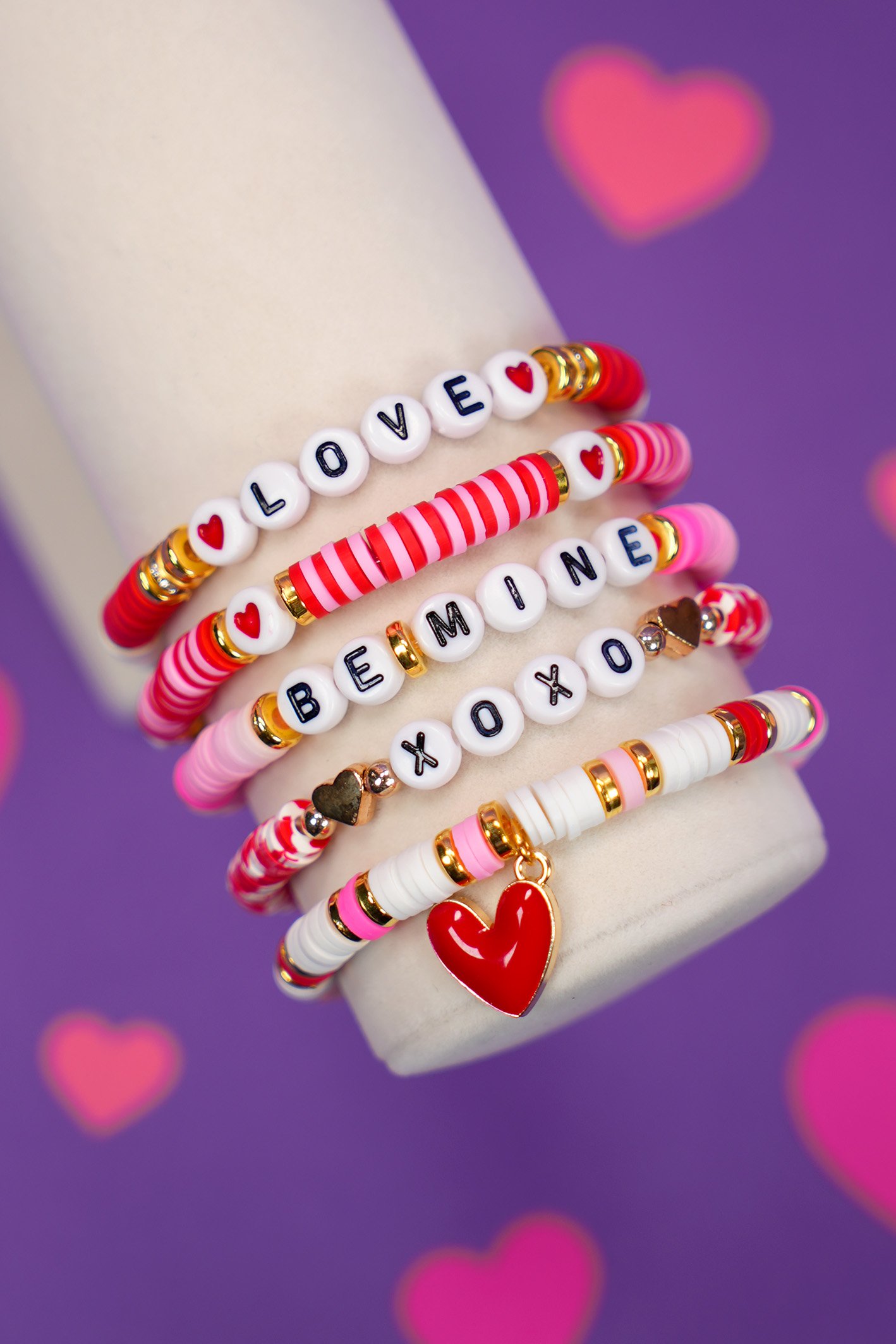 Close up of Valentine's Day bracelet set with red, pink, and white beads on display arm on purple background with pink hearts