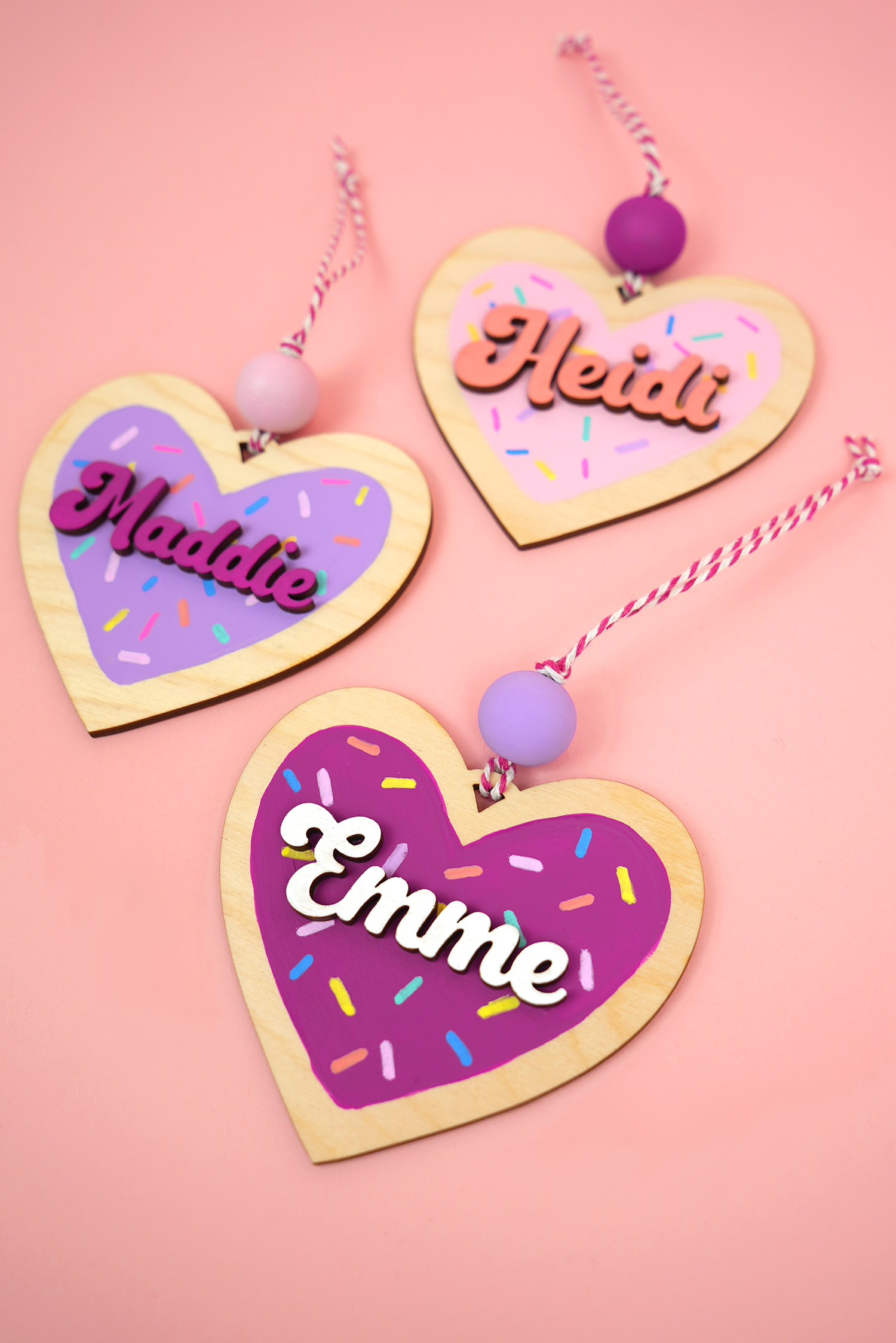 Three cute heart ornaments cut from wood and painted to look like Valentine cookies on a pink background