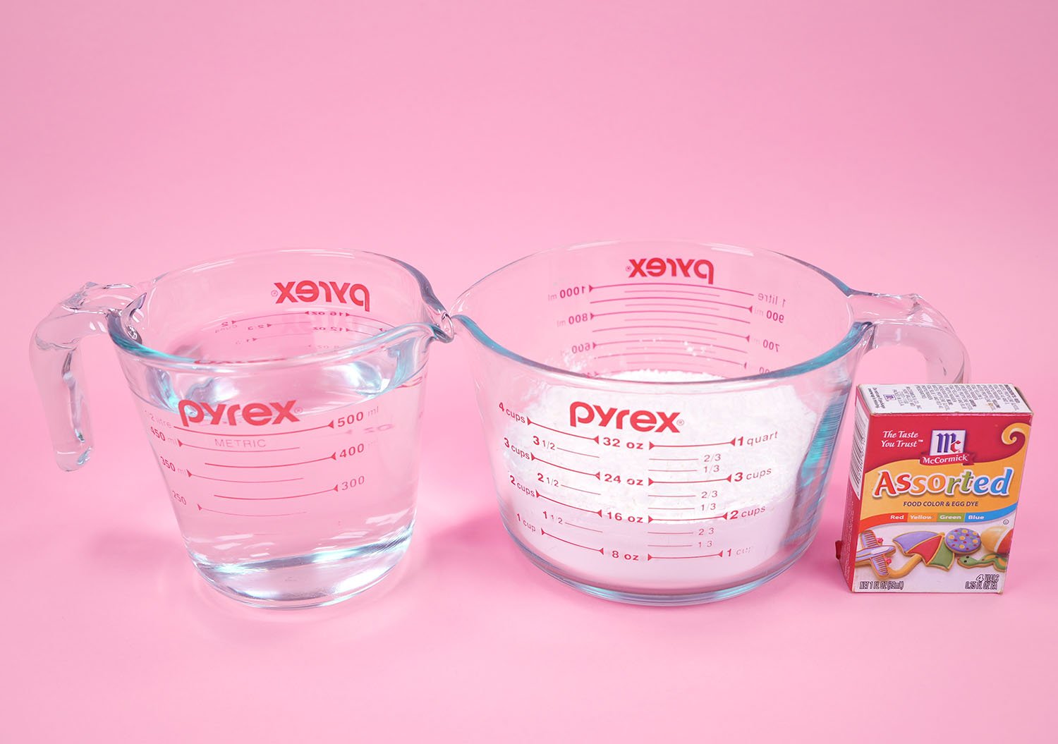 Measuring cups with cornstarch and water along with a box of food coloring on pink background